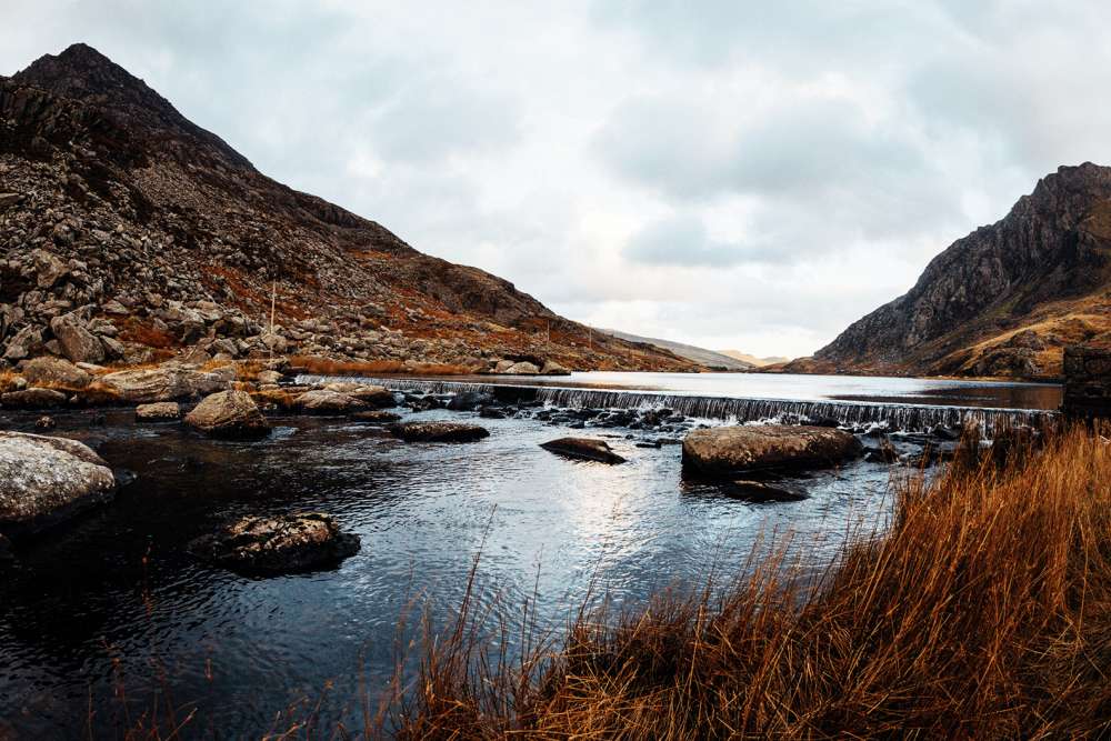 A large lake with a small waterfall, surrounded by rocks, hills, and orange grass shrubbery.